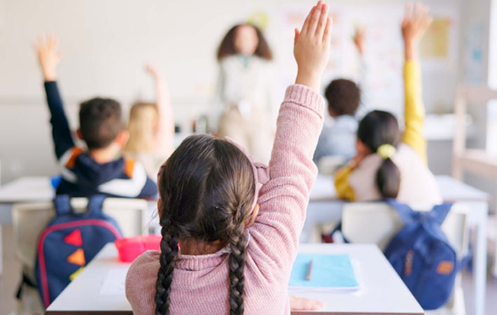 Children in a classroom raise their hands to answer a question. The teacher stands at the front of the room, out of focus. Vision and myopia control are contributors of learning in a classroom.